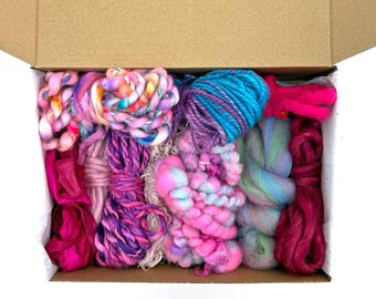 May include: A cardboard box filled with an assortment of colorful yarn and fiber materials. The collection includes various textures and shades of pink, purple, blue, and multi-colored variegated strands arranged in a dense, packed display.