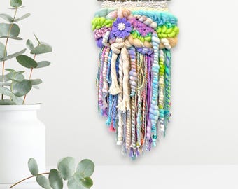 May include: A colorful fiber art wall hanging. The piece features a variety of textures and colors, including green, purple, blue, and cream. A purple flower is centered on the top. The bottom has long, dangling strands.