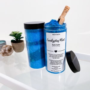 May include: A blue glass jar filled with blue bath salts and a wooden scoop. The jar has a black lid and a label that reads "RITZY BATH Eucalyptus Mint Bath Salts Net Wt. 16 oz (454g)".