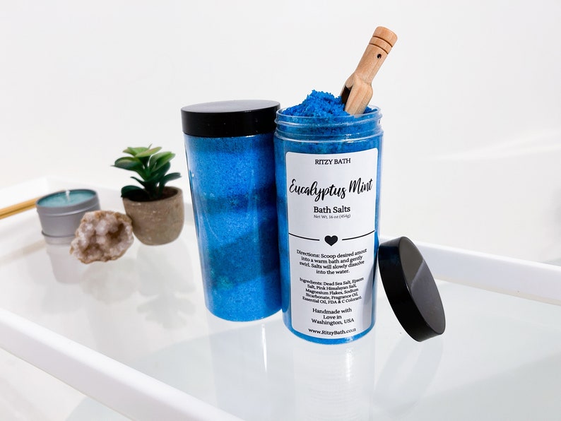 May include: A blue glass jar filled with blue bath salts and a wooden scoop. The jar has a black lid and a label that reads "RITZY BATH Eucalyptus Mint Bath Salts Net Wt. 16 oz (454g)".