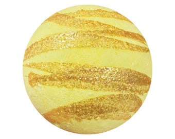 May include: A spherical bath bomb with a pale yellow base color, featuring diagonal, textured streaks of shimmering gold glitter across its surface. The object has a smooth, rounded form with a consistent, uniform texture throughout the sphere.