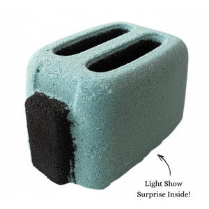 May include: A teal-colored, rectangular bath bomb shaped like a toaster. It has two rectangular slots on top and a black, textured section on the front. Text on the image reads "Light Show Surprise Inside!"