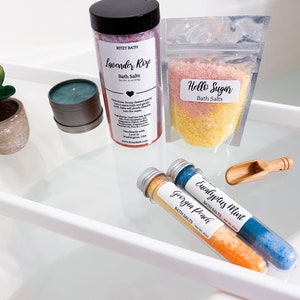 May include: Assortment of bath salts in various sizes and packaging. Lavender Rose, Hello Sugar, Eucalyptus Mint, and Georgia Peach bath salts are displayed on a white shelf.
