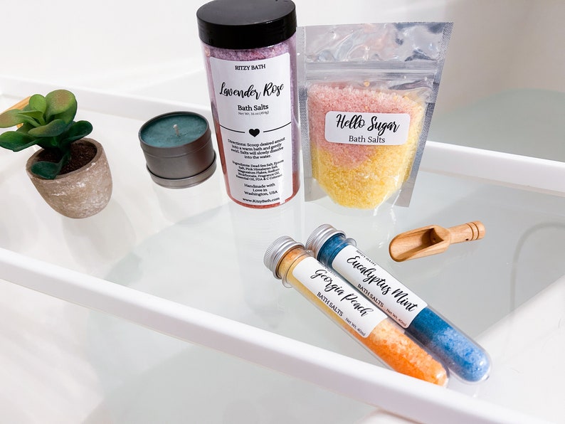 May include: Assortment of bath salts in various sizes and packaging. Lavender Rose, Hello Sugar, Eucalyptus Mint, and Georgia Peach bath salts are displayed on a white shelf.