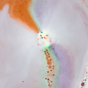 May include: Close-up of a bath bomb dissolving in water, creating a colorful, bubbly display. The colors include orange, white, purple, green, and small colorful sprinkles. The bath bomb is creating a foamy, textured surface.
