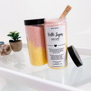 May include: Two clear plastic jars filled with pink and yellow bath salts. The jars have black lids and labels that say "Ritzy Bath" and "Hello Sugar Bath Salts".