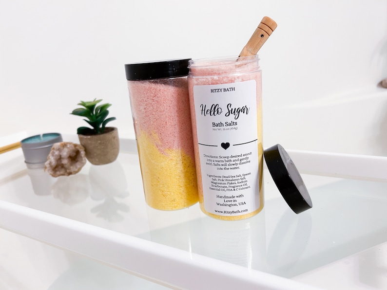 May include: Two clear plastic jars filled with pink and yellow bath salts. The jars have black lids and labels that say "Ritzy Bath" and "Hello Sugar Bath Salts".