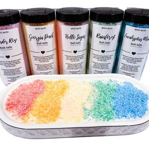 May include: Five glass jars of bath salts with colourful labels and names like Lavender Rose, Georgia Peach, Hello Sugar, Rainforest, and Eucalyptus Mint. The jars are arranged in a row on a white oval dish with a small amount of bath salts in each colour.
