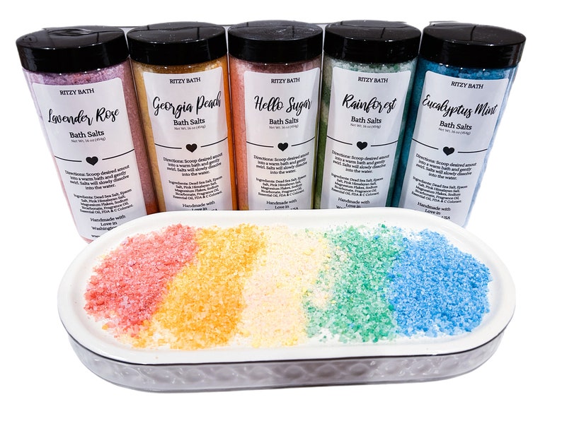May include: Five glass jars of bath salts with colourful labels and names like Lavender Rose, Georgia Peach, Hello Sugar, Rainforest, and Eucalyptus Mint. The jars are arranged in a row on a white oval dish with a small amount of bath salts in each colour.