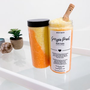 May include: Two clear glass jars filled with orange and yellow bath salts. The jars have black lids and a white label that reads "RITZY BATH Georgia Peach Bath Salts Net Wt. 16 oz (453g)". The label also includes directions for use and a list of ingredients.