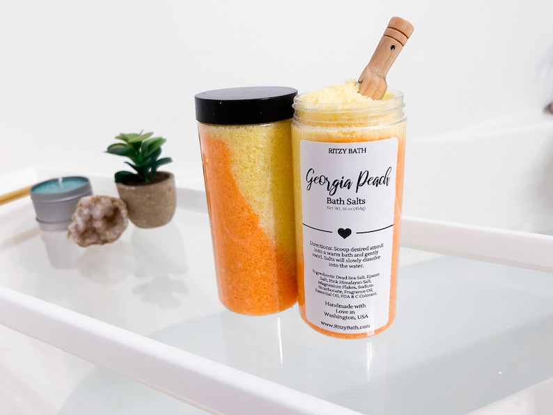 May include: Two clear glass jars filled with orange and yellow bath salts. The jars have black lids and a white label that reads "RITZY BATH Georgia Peach Bath Salts Net Wt. 16 oz (453g)". The label also includes directions for use and a list of ingredients.