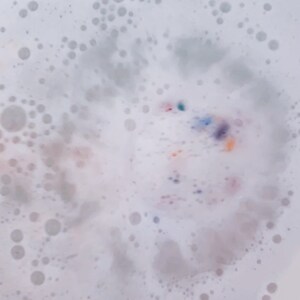 May include: Close-up of a bath with white bubbles and colorful specks. The bubbles vary in size, creating a textured surface. The colorful specks include shades of blue, purple, orange, and pink, adding visual interest to the bath water.