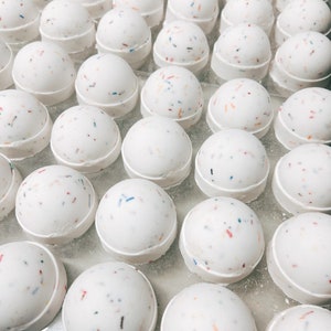 May include: A collection of white, spherical bath bombs with colorful confetti-like sprinkles. Each bath bomb sits on a white, circular base. The image is a close-up shot, showcasing the texture and design of the bath products.