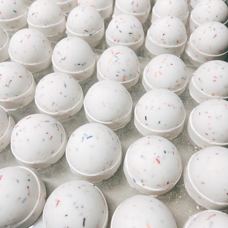 May include: A collection of white, spherical bath bombs with colorful confetti-like sprinkles. Each bath bomb sits on a white, circular base. The image is a close-up shot, showcasing the texture and design of the bath products.