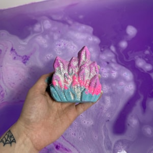 May include: A colorful bath bomb in the shape of a crystal formation. The bath bomb features pink, blue, and silver glitter accents. The bath bomb is held over a purple bath with white bubbles.