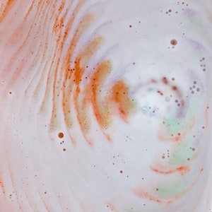 May include: Close-up of a bath bomb dissolving in water, creating a swirling pattern. The colors include white, orange, and hints of green and purple. Small bubbles dot the surface, adding texture to the image. The overall effect is a vibrant, abstract design.