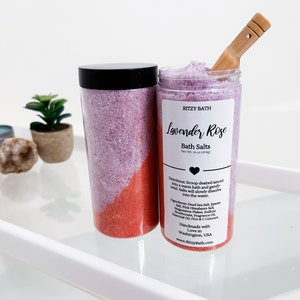 May include: A clear jar of lavender and rose bath salts with a black lid and a wooden scoop. The jar is labelled "RITZY BATH Lavender Rose Bath Salts Net Wt. 34 oz (964g)".
