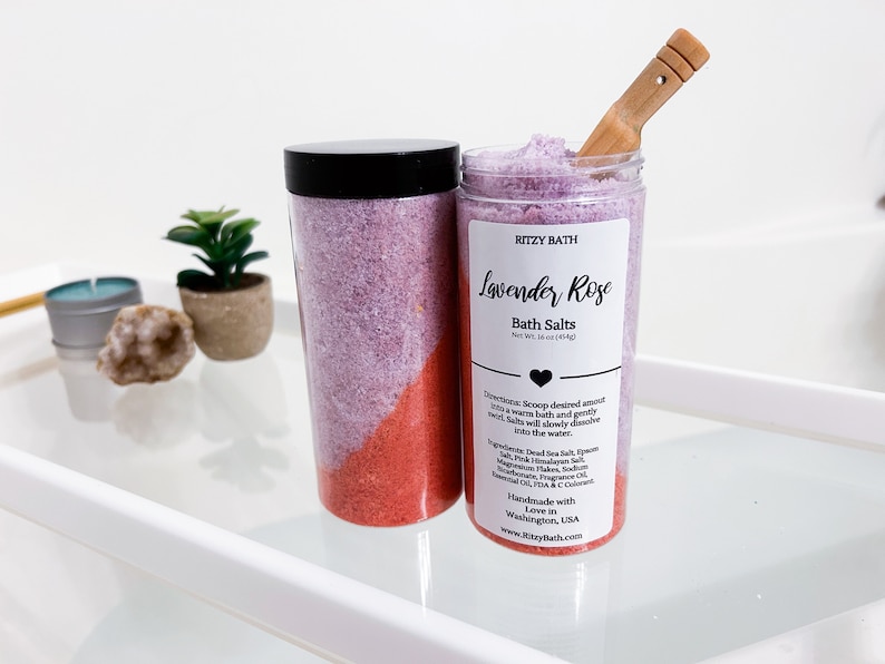 May include: A clear jar of lavender and rose bath salts with a black lid and a wooden scoop. The jar is labelled "RITZY BATH Lavender Rose Bath Salts Net Wt. 34 oz (964g)".