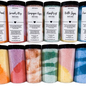 May include: Eight clear jars of bath salts with colourful layers. The jars are labelled with different scents, including Georgia Peach, Lavender Rose, Champagne Kisses, Rainforest, Hello Sugar, and Eucalyptus Mint. Each jar has a black lid.