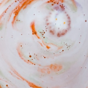 May include: Close-up of a bath bomb dissolving in water. The swirling pattern features white, orange, and brown hues, with small, scattered bubbles. The center has a lighter, almost yellow tone.