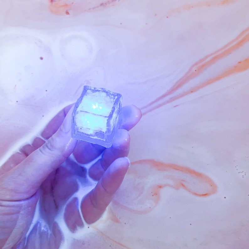 May include: A clear, cube-shaped object with a glowing blue interior is held in a hand. The background is a light pink and white liquid with orange streaks. The object appears to be a bath bomb or similar product.