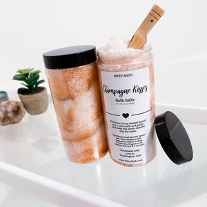 May include: A glass jar filled with pink and white bath salts with a black lid and a wooden scoop. The jar has a white label that reads "RITZY BATH Champagne Kisses Bath Salts".