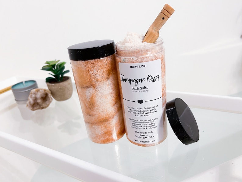 May include: A glass jar filled with pink and white bath salts with a black lid and a wooden scoop. The jar has a white label that reads "RITZY BATH Champagne Kisses Bath Salts".