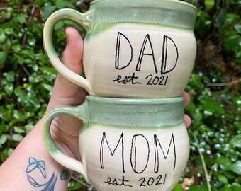 May include: Two handmade ceramic mugs, one stacked on top of the other. The top mug reads "DAD est 2021" in black script, and the bottom mug reads "MOM est 2021". Both mugs have a green rim and handle.