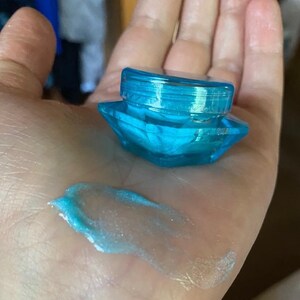 Könnte beinhalten: Ein kleines, klares blaues Glas mit einem diamantförmigen Deckel. Das Glas ist mit einem hellblauen, schimmernden Gel gefüllt.