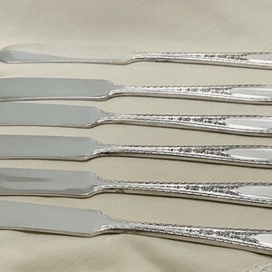 May include: Six silver butter knives with ornate handles. The knives are arranged in a stack, with the handles facing up.
