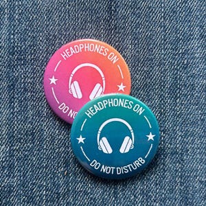 May include: Two circular badges with the text "Headphones On" and "Do Not Disturb". One is pink and orange, the other teal. Both feature a white headphone graphic and star details. The badges are on a denim background.