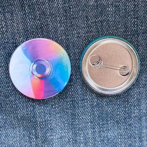 May include: Two circular pin badges. One has a holographic, rainbow design with a dark blue centre. The other shows the pinback mechanism, with a safety pin and a teal border.
