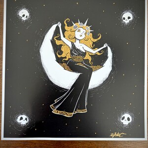 May include: Illustration of a woman with long blonde hair, wearing a black dress and a floral crown, seated on a crescent moon. The artwork features gold accents and is set against a black background with stars and skull motifs.