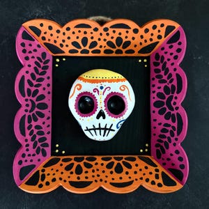 May include: A Day of the Dead themed wall hanging. The artwork features a colorful skull in a decorative frame. The frame is orange and pink with a scalloped edge and floral cutouts. The skull is white with colorful details.