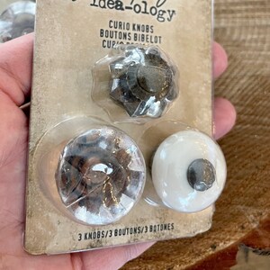May include: A package of three decorative knobs, featuring a black and silver starburst design, a brown wood-look knob, and a white knob with a silver center. The package is labeled "Jim Holtz idea-ology Curio Knobs Boutons Bibelot 3 Knobs/3 Boutons/3 Botones".