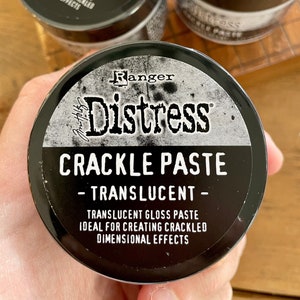 May include: A black jar of Ranger Distress Crackle Paste - Translucent. The jar has a white label with the text "Ranger Distress Crackle Paste - Translucent - Translucent Gloss Paste Ideal for Creating Crackled Dimensional Effects".
