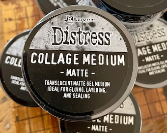 May include: Several black and silver Distress Collage Medium - Matte containers are stacked on a wooden surface. The containers have a distressed design with the product name and description in white text. The product is a translucent matte gel medium for gluing, layering, and sealing.