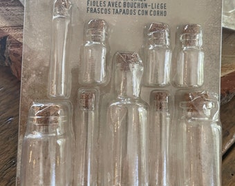 Peut inclure: Un ensemble de 9 fioles en verre bouchées dans un emballage en plastique transparent. Les fioles sont de différentes tailles et formes. L'emballage porte l'inscription "Jim Holtz idea-ology Corked Vials".
