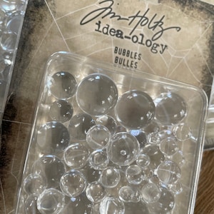 May include: A clear plastic container filled with 60 clear plastic bubbles. The container is labeled "Jim Holtz idea-ology Bubbles Bulles 60 Bubbles/60 Bulles".