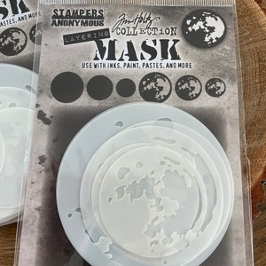 May include: A clear plastic package containing a set of four layering stencils for crafting. The stencils are white and feature a moon design. The package includes the text "Stampers Anonymous Jim Holtz Collection Layering Mask Use with inks, paint, pastes, and more."