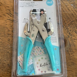 May include: A silver Crop-A-Dile hole punch and eyelet setter tool with teal handles, packaged in clear plastic. The tool has a locking system and includes the text "We R Memory Keepers" and "hello".