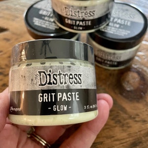May include: Three jars of Distress Grit Paste in Glow. The jars are black with white lettering and a green label. The text on the label reads "Distress Grit Paste - Glow - 3 fl oz (88 ml)"
