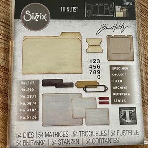 May include: A set of 54 metal dies for crafting, featuring a vintage-inspired design with a folder, labels, and numbers. The dies are packaged in a clear plastic bag with a white background and the Sizzix logo. The package also includes the text "THINLITS®" and "Jim Holtz".