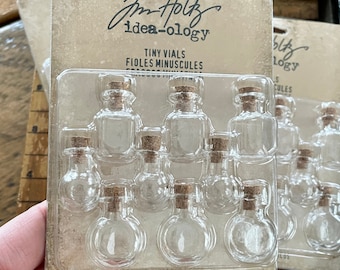 Peut inclure: Un paquet de 9 petits flacons en verre avec des bouchons en liège. L'emballage est étiqueté "Jim Holtz idea-ology Tiny Vials Fioles Minuscules 9 Vials/9 Fioles/9 Frascos".