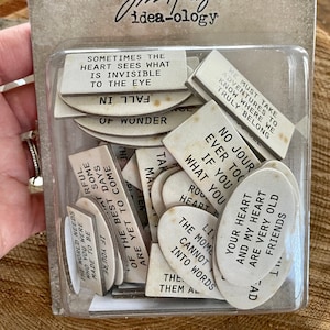 May include: A set of white wooden quote chips with various inspirational phrases, such as "Sometimes the heart sees what is invisible to the eye" and "No journey ever too far if you what you heart". The chips are packaged in a clear plastic container with a black and white label that reads "Jim Holtz idea-ology" and "Quote Chips, Copeux de Citation".