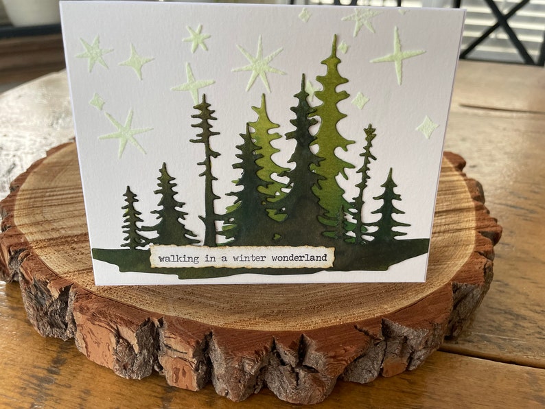 May include: A white card with a green silhouette of a forest with the text "walking in a winter wonderland" at the bottom. The background is white with green stars.