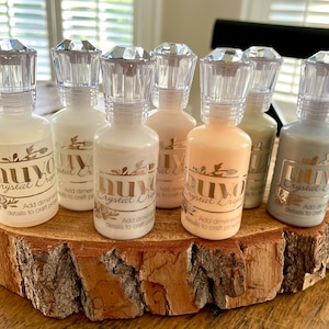 May include: Seven bottles of Nuvo Crystal Drops in various colors, including white, silver, gray, and peach. The bottles have clear diamond-shaped tops and are arranged on a wooden slice.
