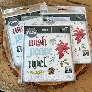 May include: Several Sizzix Thinlits die sets in clear packaging. The sets feature Christmas-themed designs, including the words "wish, peace, noel", poinsettias, and other festive elements. The packaging includes the designer's name, Jim Holtz.