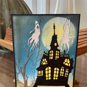 May include: A Halloween-themed card featuring a silhouette of a haunted house with glowing yellow windows and a full moon in the background. Two white ghost cutouts float above the house. The text "VANISHING VISITORS" is at the bottom.