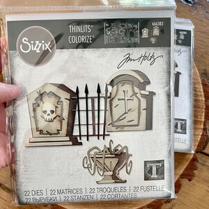 May include: A package of 22 metal dies for crafting, featuring a graveyard theme with a tombstone, gate, and urn. The dies are designed by Jin Holtz and are part of the Sizzix Thinlits Colorize collection. The package includes the text "666383" and "22 Dies | 22 Matrices | 22 Troqueles | 22 Fustelle | 22 Вырубки | 22 Stanzen | 22 Cortantes".